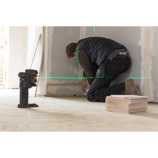 STANLEY FMHT77598-1 laserwaterpas projecteert groene lijnen voor muuruitlijning, knielende werknemer, stapel tegels zichtbaar.