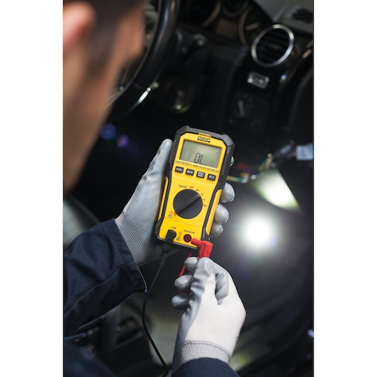 Foto van de STANLEY FMHT82563-0 digitale multimeter voor autodiagnose, vastgehouden door gehandschoende handen.