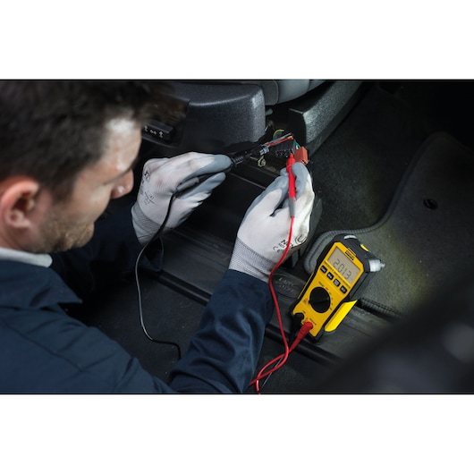 Foto van STANLEY FMHT82563-0 digitale multimeter bij het testen van autokabels, gele behuizing, duidelijk display.