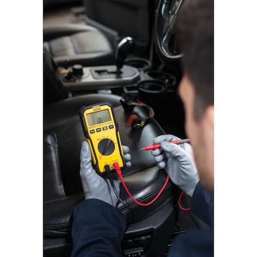Foto van STANLEY FMHT82563-0 digitale multimeter in gebruik binnen een auto, met gehandschoende handen en meetpennen.