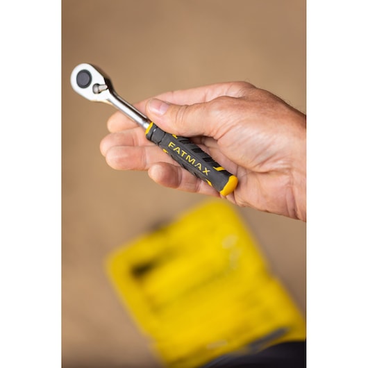 Foto van een hand met een STANLEY FATMAX ratel, SKU FMMT82683-0, zwart-geel handvat, onscherpe gele gereedschapskoffer eronder.