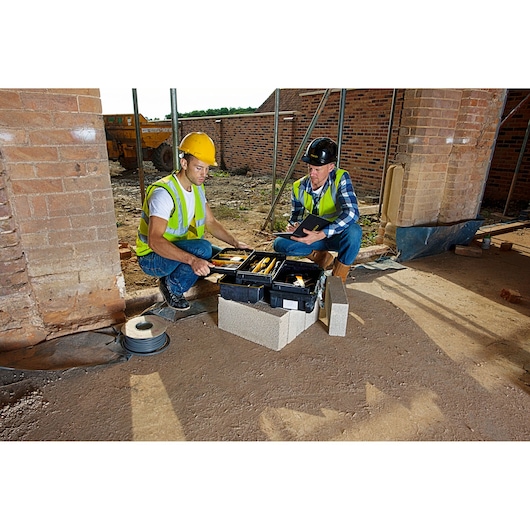 Boîte à outils STANLEY FMST1-71219 ouverte sur chantier, montrant des outils à main utilisés par deux ouvriers.