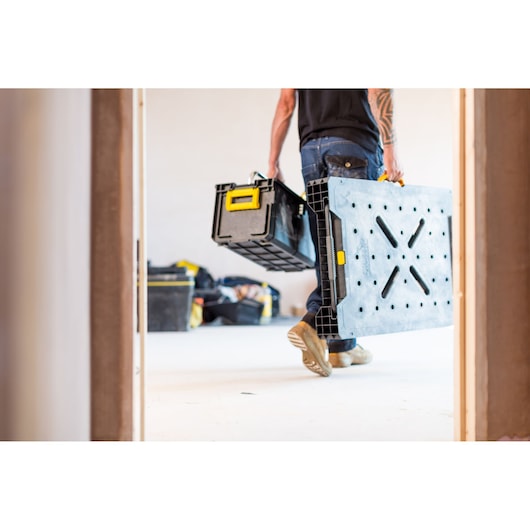 STANLEY FMST1-75672 draagbare opvouwbare werkbank gedragen door persoon, zwart met gele accenten, binnenruimte.