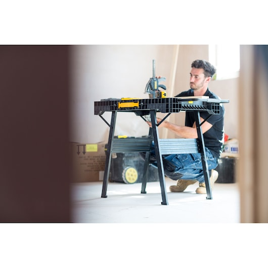 Foto van STANLEY FMST1-75672 werkbank in gebruik op bouwplaats, persoon knielt naast zwarte opvouwbare tafel.