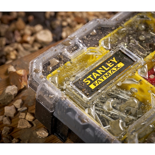 Photo du rangement étanche pour petites pièces STANLEY FMST1-75781 avec gouttelettes d'eau sur le couvercle, entouré de pierres.