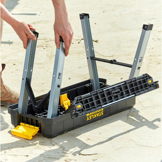 STANLEY FMST81083-1 opvouwbare werkbank wordt buiten gemonteerd, met zwarte en gele kunststof onderdelen en metalen poten.