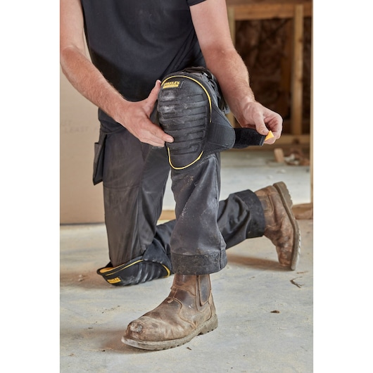 STANLEY FMST82959-1 kniebeschermer met zwarte en gele rand wordt bevestigd om werkbroek, bouwomgeving.