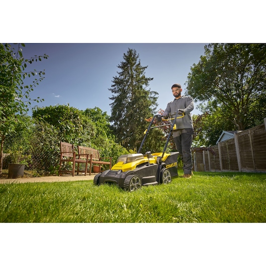 Photo de la tondeuse STANLEY SCMWP3701M2 en utilisation sur pelouse dans un jardin, design jaune et noir visible.