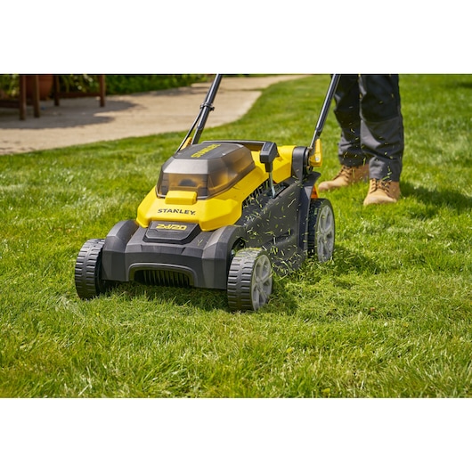 Tondeuse à gazon STANLEY SCMWP3701N jaune et noire, utilisée sur de l’herbe, avec des coupeaux visibles et jambes de l’utilisateur.