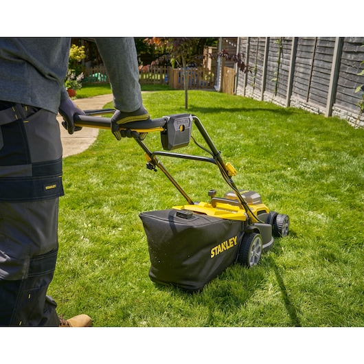 Photo de la tondeuse sans fil STANLEY SCMWP3701N utilisée sur une pelouse, angle arrière montrant le bac de ramassage.