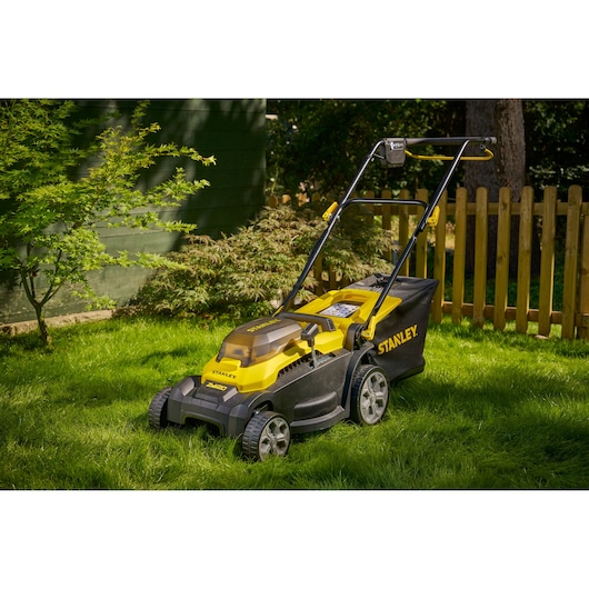 Tondeuse STANLEY SCMWP3701N jaune et noire sur herbe, clôture et feuillage en arrière-plan.