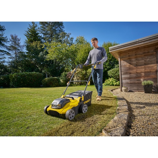 Foto van STANLEY SCOEP3801M2 gazonverticuteermachine in gebruik op gras bij een houten schuur, door persoon geduwd.