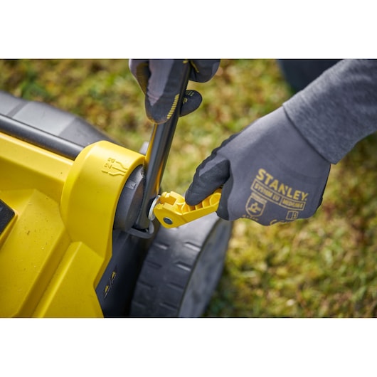 STANLEY SCOEP3801N fotografie van een gehandschoende hand die gele grasmaaierhendel buiten afstelt.
