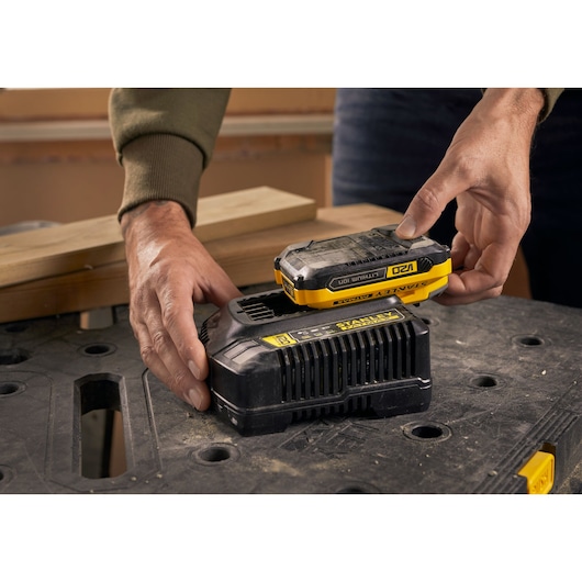 Foto van STANLEY SFMCB14 batterijoplader met batterij op werkbank, handen plaatsen batterij op oplader.