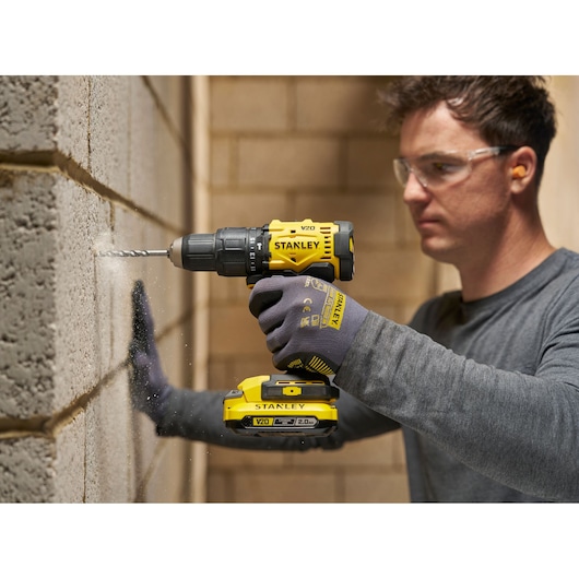 Persoon boort in een betonnen muur met een gele STANLEY accuboormachine, draagt handschoenen en bril.