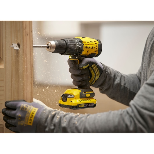 Persoon boort een gat in hout met een Stanley V20 accuboormachine, draagt handschoenen en maakt houtspaanders.