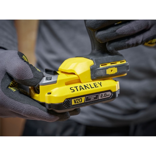 Close-up van handen met handschoenen die een gele STANLEY V20 2.0Ah lithium-ion batterij vasthouden.