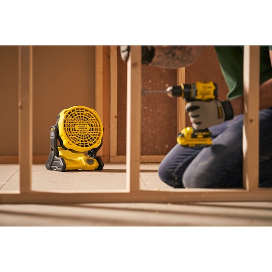 Ventilateur de chantier portable Stanley SFMCE001B au sol dans un chantier, ouvrier utilisant une perceuse à proximité.
