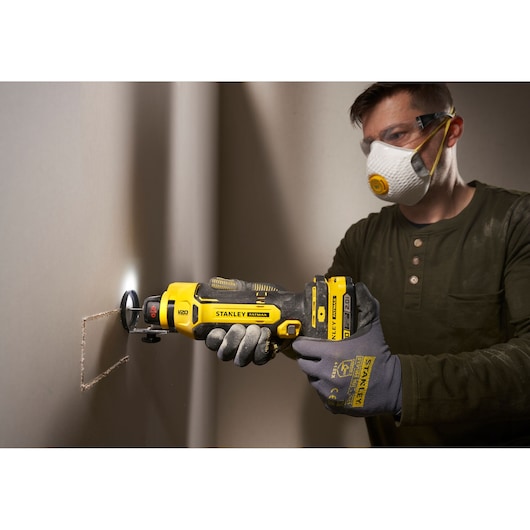 Foto van STANLEY SFMCE200B roterend gereedschap dat gipsplaat snijdt, vastgehouden door handschoenen, op de werkplek.