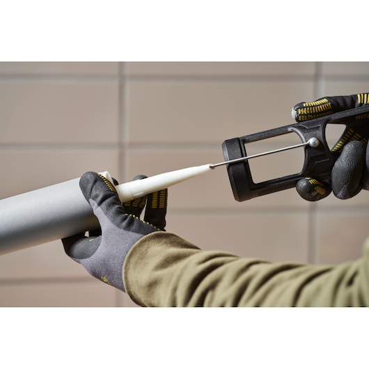 Photo du pistolet à calfeutrer STANLEY SFMCE600B tenu par des mains gantées, application de mastic, vue latérale.