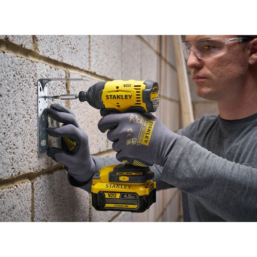 Stanley V20 visseuse à choc sans fil fixant un support métallique au mur, utilisateur avec gants et lunettes.