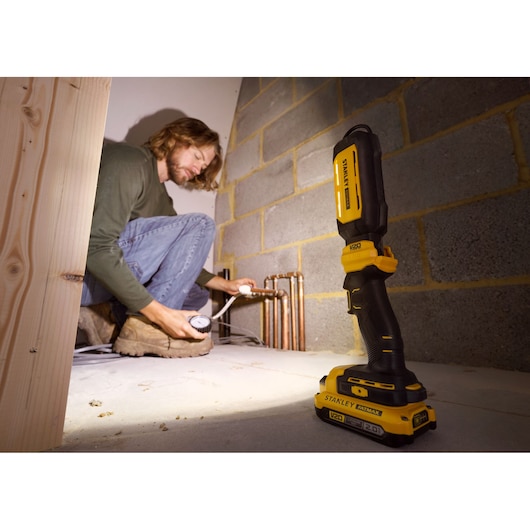 STANLEY SFMCL050B 18V FATMAX V20 Pivot Light op de vloer, verlicht leidingen, persoon werkt op achtergrond.