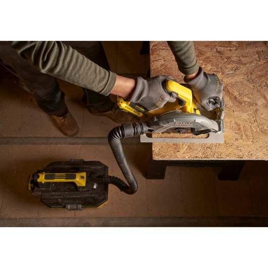 Vue du dessus d'une personne utilisant une scie circulaire sans fil STANLEY SFMCS550B pour couper du bois, reliée à un extracteur de poussière.