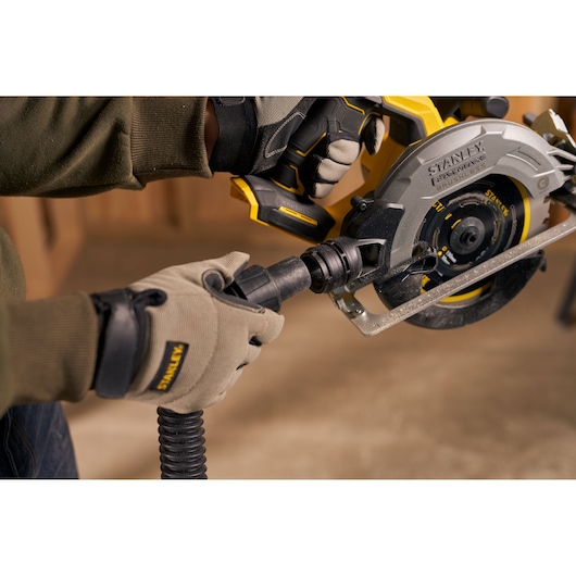 Scie circulaire STANLEY SFMCS550B avec raccord d’aspirateur tenue par des mains gantées en atelier.