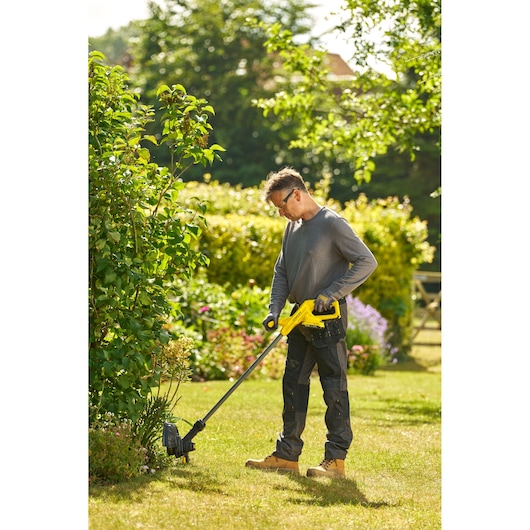 Photo d'une personne utilisant un coupe-bordures STANLEY SFMCST925B dans un jardin, taillant l'herbe près des buissons.