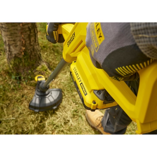 Photographie du coupe-herbe jaune STANLEY SFMCST925B utilisé près d'un tronc d'arbre sur de l'herbe.