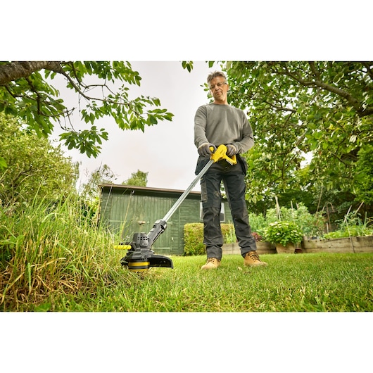 STANLEY SFMCST925M1 coupe-bordures en utilisation, personne coupant la pelouse dans un jardin avec abri vert en arrière-plan.