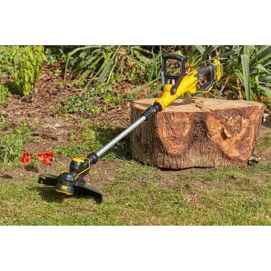 STANLEY SFMCST933B draadloze grastrimmer in geel en zwart op gras bij een boomstronk buiten.