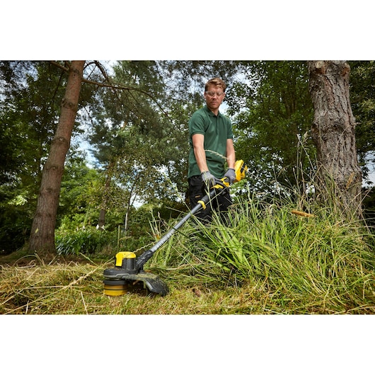 STANLEY SFMCSTB963B grastrimmer in gebruik buiten voor het maaien van hoog gras, bediend door iemand met tuinhandschoenen.