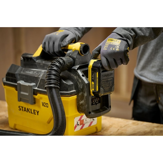 Stanley V20 draadloze stofzuiger, persoon met handschoenen plaatst batterij op geel-zwarte machine.