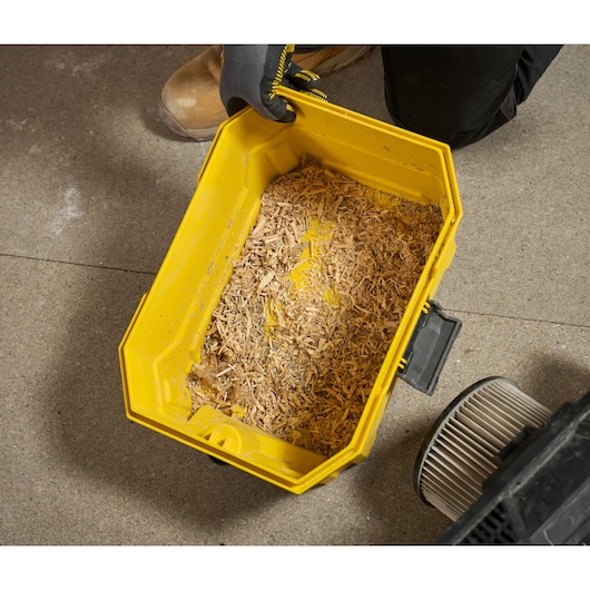 Gele stofzuigerbak met houtspaanders op werkplaatsvloer, filter en een hand met handschoen zichtbaar.
