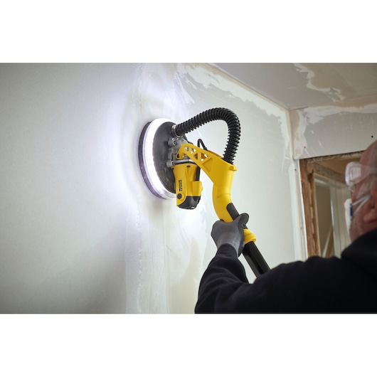 STANLEY SFMEE500S gipswand schuurmachine in gebruik, gele handgreep, schuurt witte muur, close-up.
