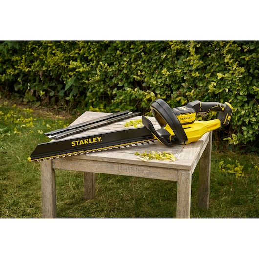 STANLEY STCHT8255B gele en zwarte heggenschaar op houten tafel buiten, bladen en bladeren zichtbaar.