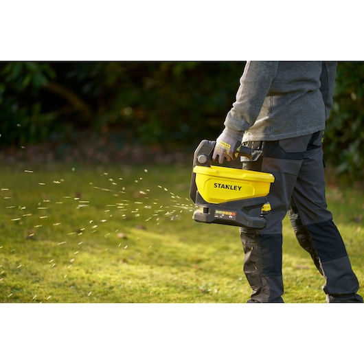 Photographie d'une personne utilisant l’épandeur manuel STANLEY STCOE300B jaune et noir, dispersant du matériau sur l’herbe.
