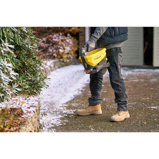 Photo de l’épandeur manuel STANLEY STCOE300B utilisé sur un sentier de jardin enneigé, design jaune et noir.