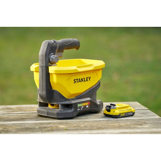 STANLEY STCOE300B épandeur jaune portable et batterie sur surface en bois extérieure, vue latérale.