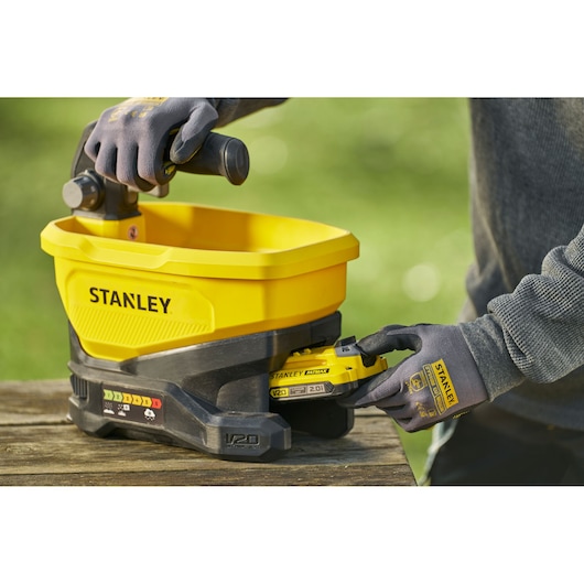 Stanley STCOE300B épandeur de jardin jaune sans fil avec batterie V20, vue latérale, sur table en bois.