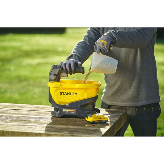 STANLEY STCOE300B épandeur sur table avec trémie jaune, batterie fixée, personne versant du matériau.