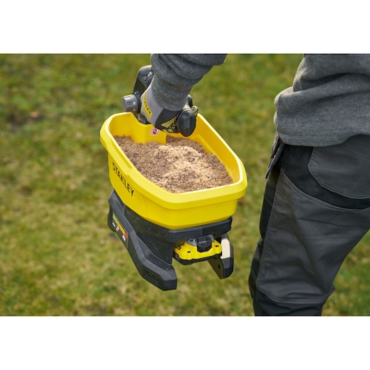 Stanley STCOE300B épandeur portable avec bac jaune rempli de sable, tenu par une personne en extérieur.