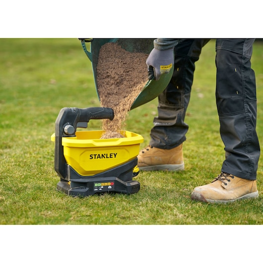 Stanley STCOE300B épandeur, posé sur l'herbe, rempli de sable par une personne avec des gants et des bottes de travail.