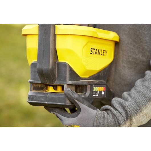 STANLEY STCOE300B épandeur jaune tenu par une main gantée, vue latérale, fond extérieur.