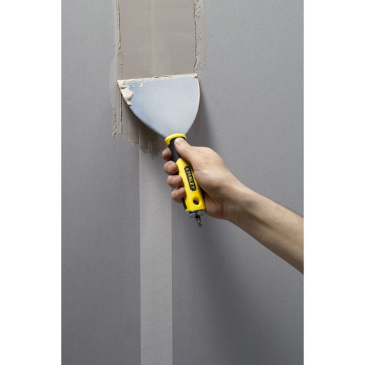 STANLEY STHT0-28034 plamuurmes met geel-zwarte handgreep brengt vulmiddel aan op een muur.