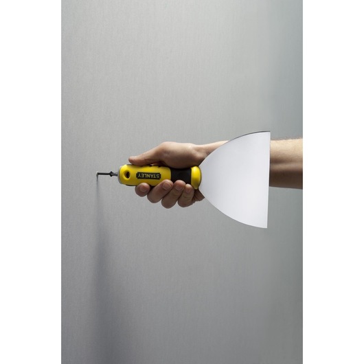 STANLEY STHT0-28034 plamuurmes met geel-zwarte handgreep, zijaanzicht vastgehouden door hand tegen muur.