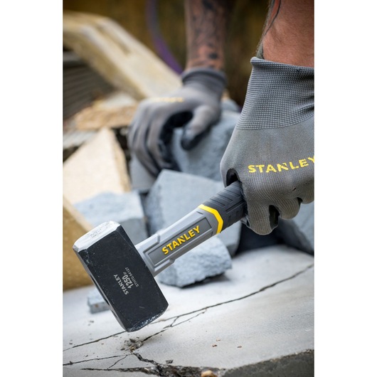 Photo du marteau STANLEY STHT0-54127 en utilisation, tête noire avec accents jaunes, tenu par une main gantée sur du béton.