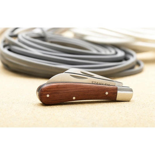 STANLEY STHT0-62687 zakmes met houten handvat en ingeklapte lemmet op beige oppervlak, opgerolde kabels op de achtergrond.