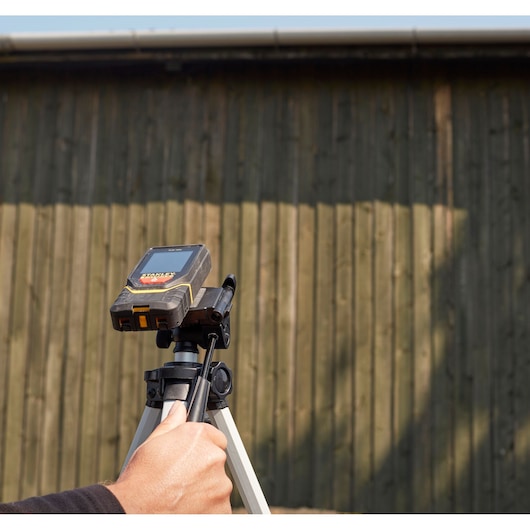 STANLEY STHT1-77139 laser afstandsmeter op statief, vastgehouden door hand, buiten met houten muur.